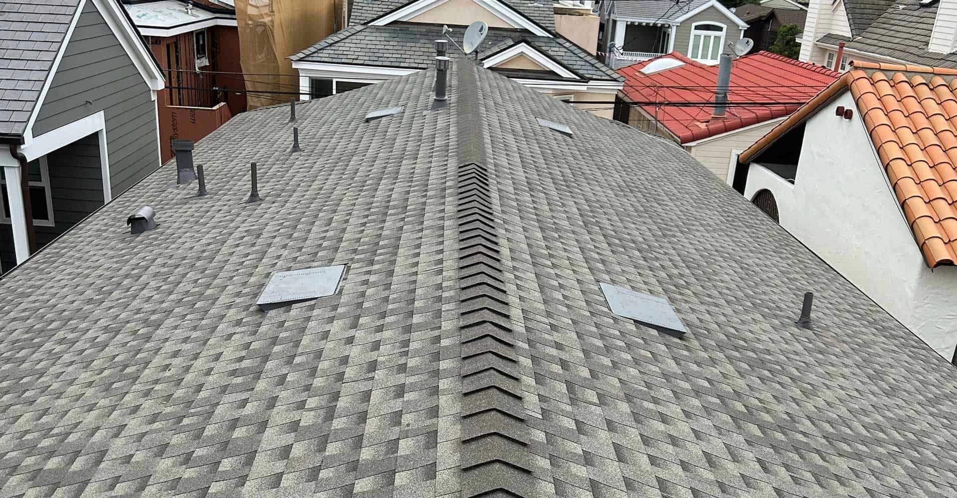 Aerial view of a gray shingled roof with several vents, possibly inspected by a roofing contractor. The roof's peak runs diagonally through the center of the image. Adjacent homes with varied roofs are visible in the background, hinting at diverse architectural styles and potential roof replacements.