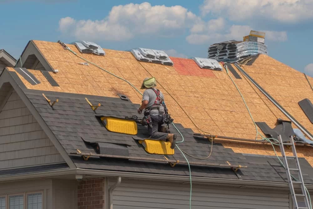 A construction worker is installing shingles on a house roof. The roof is partially covered with wooden boards and bundles of shingles. The worker is wearing safety gear and is attached to a harness. A clear sky is in the background.