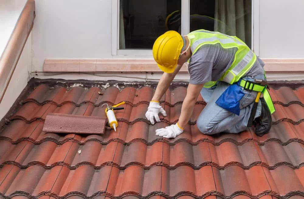 Worker in a yellow hard hat and safety vest repairing a red tile roof. They are kneeling, using tools, with a few tiles and a tube of sealant next to them. A window is visible in the background.