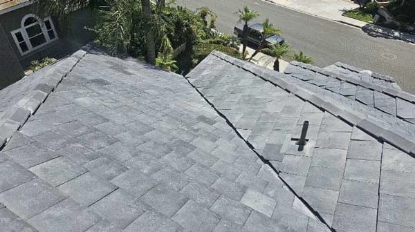 Aerial view of a residential rooftop covered with gray shingles, showcasing the need for roof maintenance. Nearby, trees loom over a parked car on the street. A roof vent is visible on the right side, hinting at recent work by a roofing contractor.