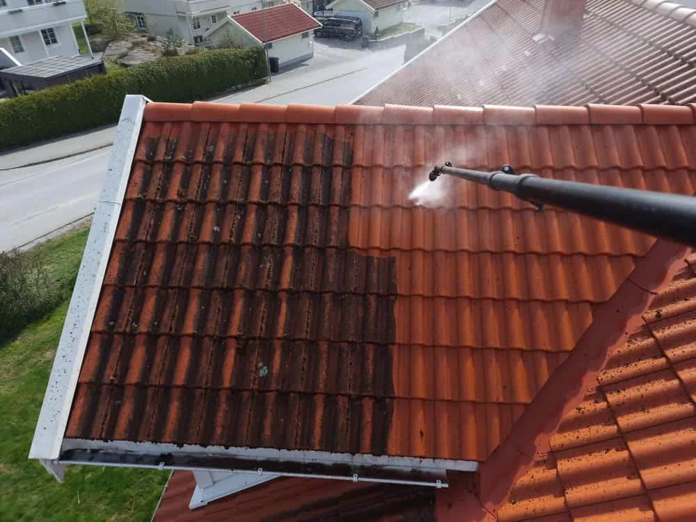 A person uses a pressure washer to clean dark, mossy tiles on a sloped roof, revealing clean, orange tiles underneath. The street and nearby houses are visible in the background.